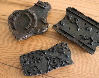 May include: Three antique, dark-colored wooden printing blocks with carved designs. One block features a circular pattern with a toothed edge, while another has a rectangular shape with floral motifs. The third block has a pattern of dots and swirls.