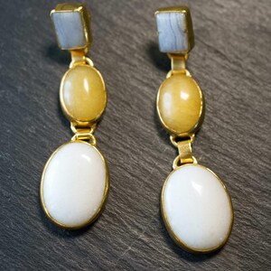 Può includere: Un paio di orecchini pendenti dorati. Ogni orecchino presenta una pietra quadrata azzurra in alto, seguita da una pietra ovale gialla e una grande pietra ovale bianca in basso. Le pietre sono incastonate in metallo dorato.