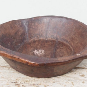 Puede incluir: Un tazón grande de madera marrón oscuro con forma redondeada y un borde ligeramente elevado. El tazón tiene dos pequeñas asas a cada lado.