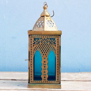 May include: A gold metal lantern with intricate cutouts and a blue glass panel. The lantern has a hinged door and a hook on the top.