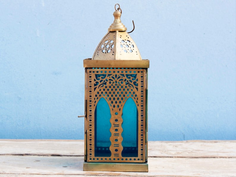 May include: A gold metal lantern with intricate cutouts and a blue glass panel. The lantern has a hinged door and a hook on the top.