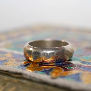 May include: A silver ring with a hammered texture rests on a colourful, patterned surface. The ring has a wide band and a slightly rounded shape. The background features blues, greens, and oranges.