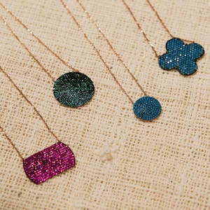 Może przedstawiać: Pięć naszyjników z różnymi kształtami i kolorami zawieszek. Zawieszki wykonane są z małych, błyszczących kamieni. Naszyjniki leżą na jasnobrązowym tle z tkaniny.