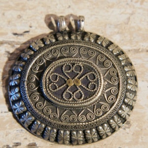 Może przedstawiać: Srebrny wisiorek z misternymi wzorami filigranowymi. Wisiorek ma centralny owal z niebieskim tłem i złotym akcentem. Wisiorek jest otoczony okrągłą obwódką z powtarzającym się wzorem małych kwiatów.