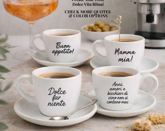 May include: Four white ceramic espresso cups with saucers, each featuring black Italian text. The cups display the phrases Buon appetito!, Mamma mia!, Dolce far niente, and Anni, amori e bicchieri di vino non si contano mai. A silver spoon rests on a saucer.