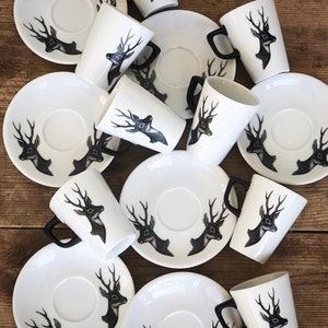 May include: White ceramic mugs and saucers with a black deer head design. The mugs have black handles. The saucers and mugs are arranged on a wooden surface. The deer head design is repeated on both the mugs and saucers.