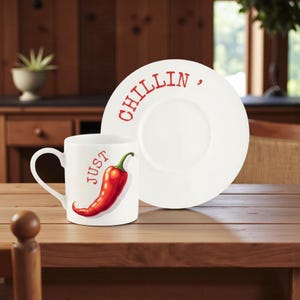Può includere: Un set di tazza e piatto in ceramica bianca. La tazza presenta un'illustrazione di peperoncino rosso con la parola "JUST" stampata verticalmente. Il piatto ha la parola "CHILLIN" in rosso. Il set è su un tavolo di legno.