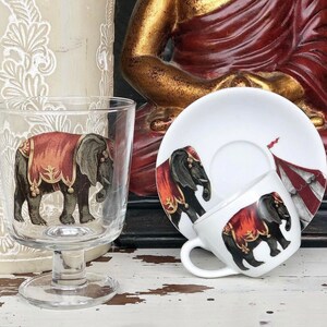 Puede incluir: Una taza de té y un platillo de cerámica blanca con un diseño de elefante rojo y dorado. El platillo tiene un diseño de carpa a rayas rojas y blancas. La taza de té y el platillo están sobre una superficie de madera blanca.