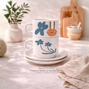 Pode incluir: Duas canecas de cerâmica brancas empilhadas em um pires branco. A caneca superior tem um design de flor azul e coelho laranja. A caneca inferior tem um design de flor azul. A imagem inclui o texto: "Opções disponíveis: Espresso (80ml) ou Chá (160ml) Tamanho Com ou sem pires."