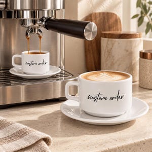 Puede incluir: Dos tazas y platillos de cerámica blanca con el texto custom order impreso en letra cursiva negra. Una taza está colocada bajo una máquina de café espresso de acero inoxidable, mientras la otra contiene café con arte latte.