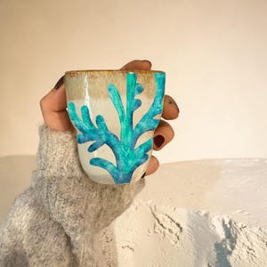 May include: A ceramic cup with a white base and a teal coral design. The rim is a light brown color. The cup is held by a person wearing a gray sweater.