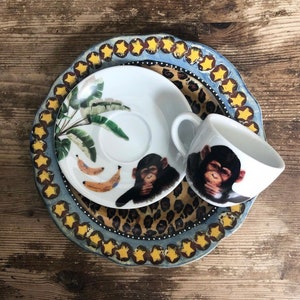 May include: A white ceramic teacup and saucer with a jungle theme. The saucer features a green palm tree and yellow bananas. The teacup has a black and white chimpanzee with its hands over its mouth.