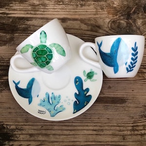 May include: Two white ceramic coffee cups with saucers. One cup features a green sea turtle with a blue whale on the saucer. The other cup features a blue whale with a green sea turtle on the saucer.