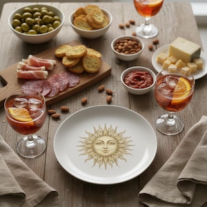 Puede incluir: Una mesa de madera con aperitivos y bebidas. Incluye aceitunas, galletas saladas, embutidos, queso y un plato con un diseño de sol. Se ven dos copas de cócteles con adorno de naranja.
