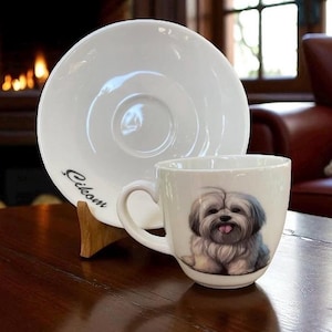 May include: White ceramic teacup and saucer set. The teacup features a detailed illustration of a fluffy, gray and white dog with its tongue out. The saucer has the word "Cikom" printed on it.