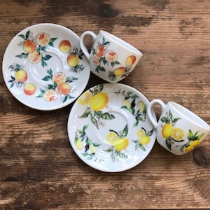Pode incluir: Duas chávenas de chá de cerâmica branca e pires com um design de limão e flores. As chávenas e os pires têm um fundo branco com limões amarelos, folhas verdes e flores brancas.