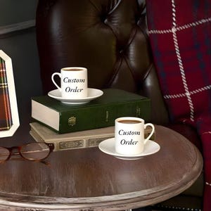 May include: Two small white coffee cups with saucers, filled with coffee, are on a wooden table. The cups have the words "Custom Order" printed on them. A stack of books and a pair of spectacles are also on the table.