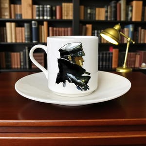 Könnte beinhalten: Weißes Keramik-Kaffeebecher- und Untertassen-Set. Die Tasse zeigt eine Aquarellillustration einer Person in einem schwarzen Mantel und Hut. Die Tasse steht auf einer weißen Untertasse, die auf einer dunklen Holzoberfläche steht.