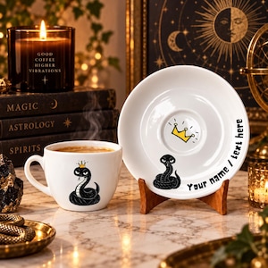 Puede incluir: Un juego de taza de café y platillo de cerámica blanca con una ilustración de una serpiente negra con una corona amarilla. La taza muestra la serpiente, mientras que el platillo presenta una serpiente, una corona amarilla y el texto Your name / text here.