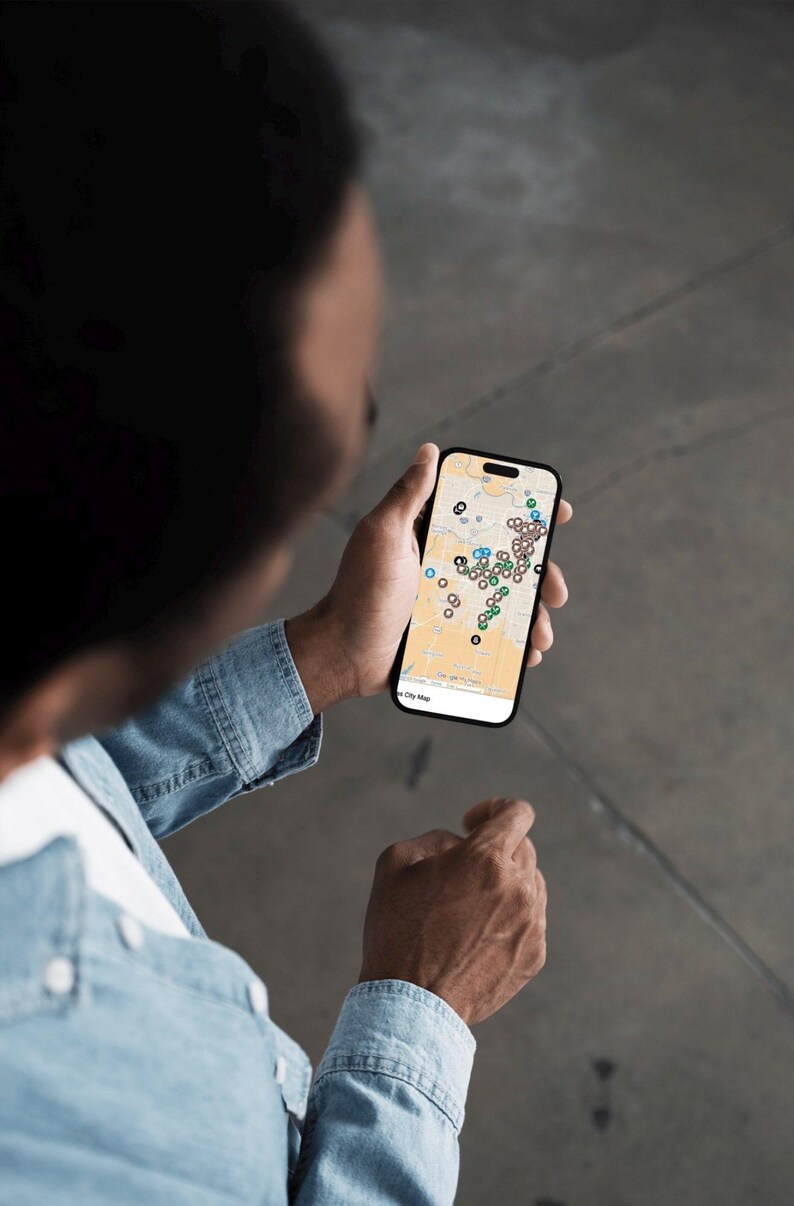 Puede incluir: Una persona sostiene un tel&eacute;fono inteligente que muestra un mapa de la ciudad con varios marcadores de ubicaci&oacute;n de colores. El tel&eacute;fono tiene un marco negro y una pantalla blanca. La persona lleva una camisa vaquera.