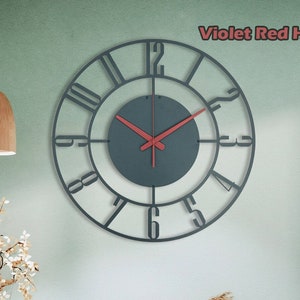 May include: A black metal clock with Roman numerals and red hands hanging on a light green wall. The clock is in a living room setting with a white chair, a small table, and plants.