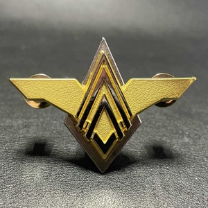 Op de afbeelding: Een goud- en zilverkleurige metalen pin met een geometrisch ontwerp. De pin heeft een centrale ruitvorm met een gelaagd, hoekig ontwerp en twee gestileerde vleugels. Zwarte accenten voegen contrast toe aan de metalen oppervlakken.