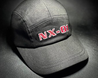May include: A black baseball cap with a curved brim. The cap features the embroidered text "NX-01" in red on the front. The cap is made of a textured fabric and has a structured design.