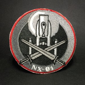 Puede incluir: Un parche bordado en plata y rojo con una nave espacial y espadas cruzadas. El texto "NX-01" está impreso en el parche.