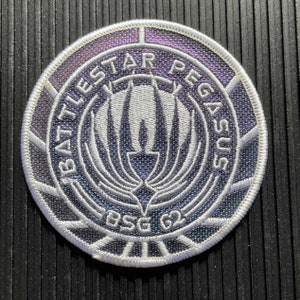 Peut inclure: Un écusson brodé blanc sur fond argenté. L'écusson présente les mots "Battlestar Pegasus" et "BSC 62" dans un design circulaire. L'écusson a un effet holographique.