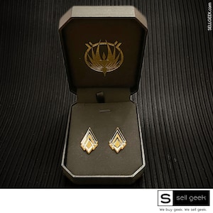 Op de afbeelding: Een paar geometrische oorbellen in goud en zwart in een zwarte fluwelen juwelendoos. De doos heeft een gouden logo met een gestileerd vogelontwerp.