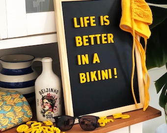 Può includere: Una lavagna nera con lettere gialle che compongono la frase "Life is better in a bikini!" La lavagna è su uno scaffale di legno con un cappello giallo, una bottiglia di liquore e un paio di occhiali da sole.