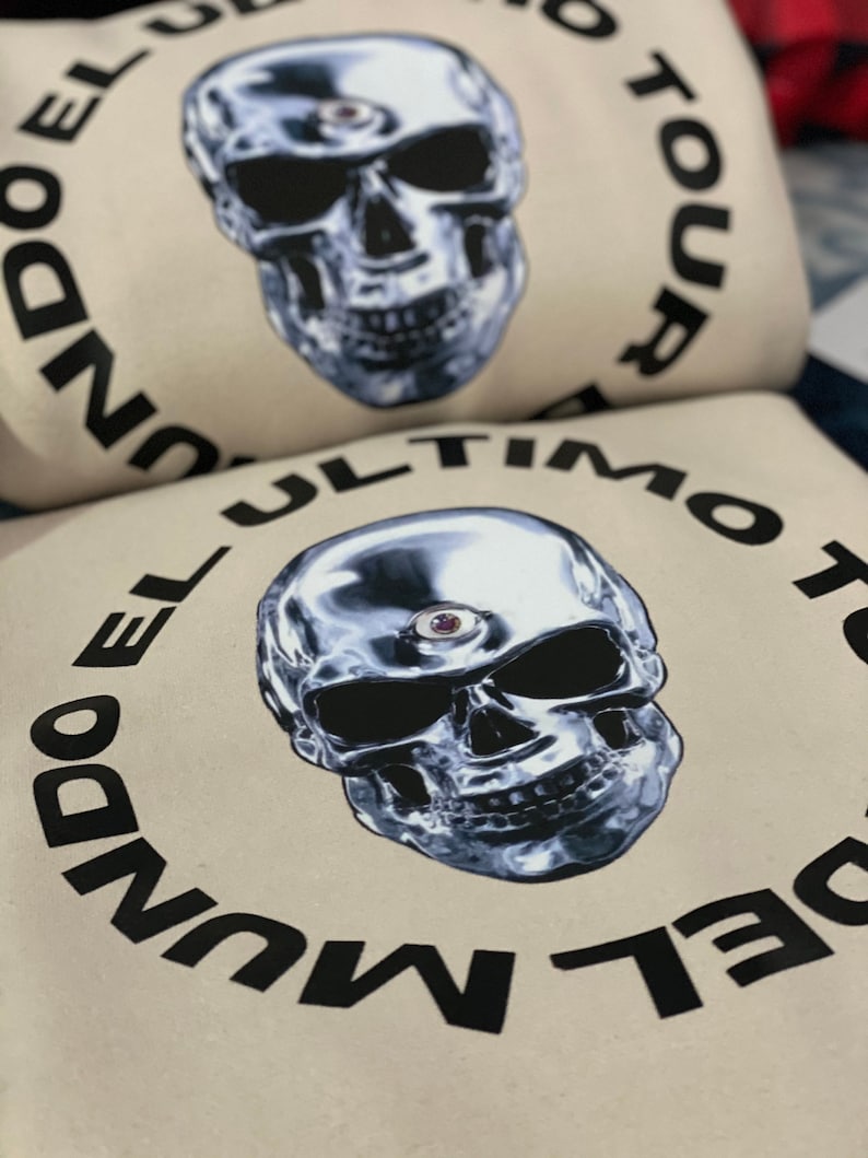 May include: Off-white sweatshirt featuring a metallic skull graphic with a third eye. The text "EL ULTIMO TOUR DEL MUNDO" is printed in a circular pattern around the skull. The design is in black and silver.
