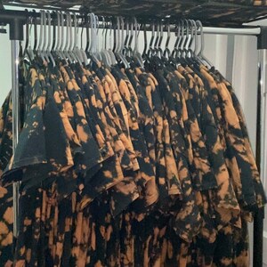 May include: A rack of black and brown tie-dye t-shirts hanging on hangers. The shirts have a unique, abstract pattern.