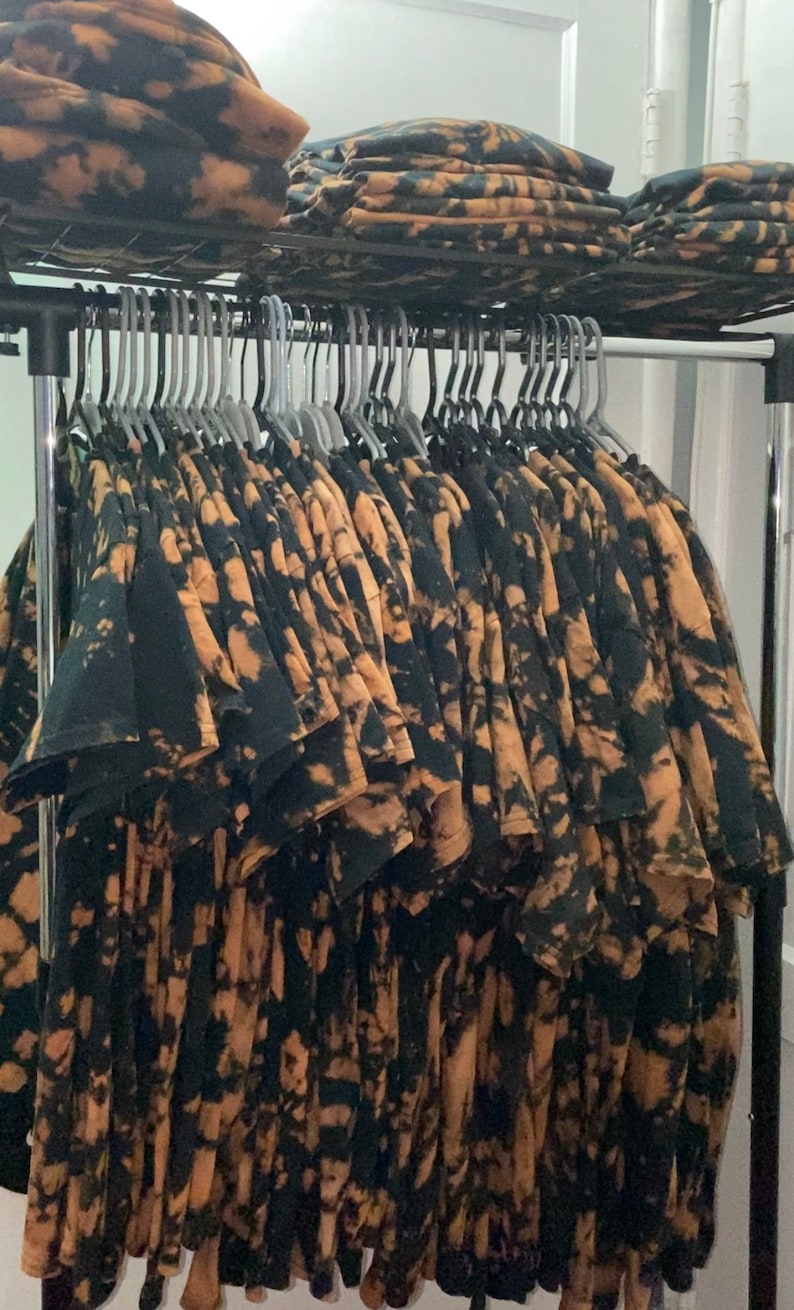 May include: A rack of black and brown tie-dye t-shirts hanging on hangers. The shirts have a unique, abstract pattern.