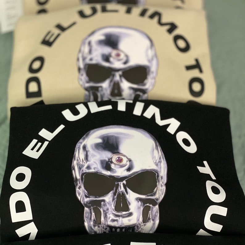 May include: Stacked sweatshirts with a metallic skull graphic and the text "DO EL ULTIMO TOUR" in a circular design. The sweatshirts are in black and beige colors.