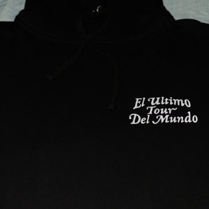 May include: Black hooded sweatshirt with white text that reads "El Ultimo Tour Del Mundo". The hoodie has a drawstring and a hood. The sweatshirt is made of a soft material.