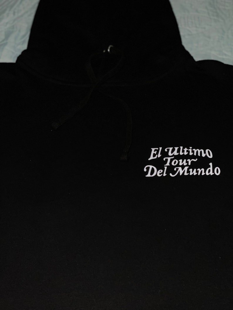May include: Black hooded sweatshirt with white text that reads "El Ultimo Tour Del Mundo". The hoodie has a drawstring and a hood. The sweatshirt is made of a soft material.