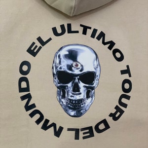May include: Beige hoodie with a metallic skull graphic in a black circular frame. The skull has a third eye and is surrounded by the text "EL ULTIMO TOUR DEL MUNDO". The hoodie's hood is visible at the top.