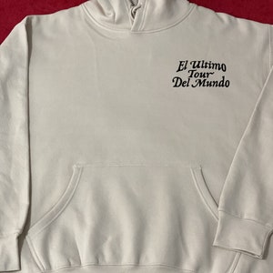 May include: A light beige hoodie with a front pocket and a hood. The text "El Ultimo Tour Del Mundo" is printed in black on the upper back. The hoodie has long sleeves and a relaxed fit.