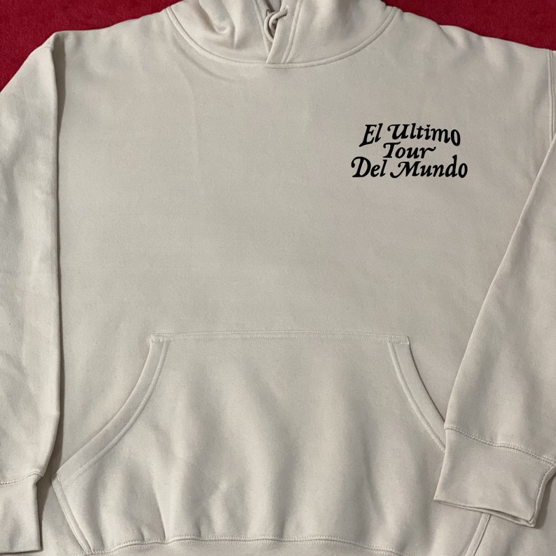 May include: A light beige hoodie with a front pocket and a hood. The text "El Ultimo Tour Del Mundo" is printed in black on the upper back. The hoodie has long sleeves and a relaxed fit.