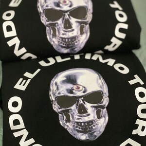 May include: Black sweatshirt featuring a metallic skull graphic with a third eye. The skull is surrounded by the white text "EL ULTIMO TOUR DEL MUNDO". The design is centered on the front of the sweatshirt.
