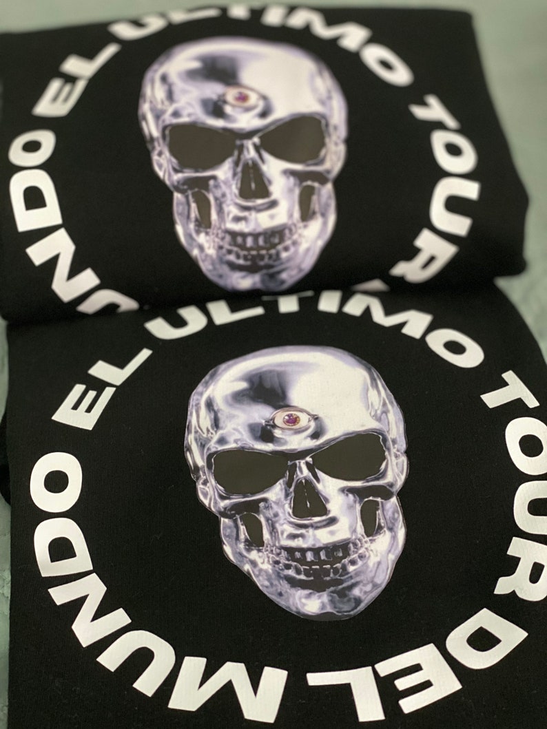 May include: Black sweatshirt featuring a metallic skull graphic with a third eye. The skull is surrounded by the white text "EL ULTIMO TOUR DEL MUNDO". The design is centered on the front of the sweatshirt.