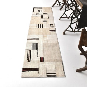 Pode incluir: Um tapete corredor branco e creme com um padrão geométrico de listras pretas e marrons. O tapete é feito de tecido tecido e tem um design de patchwork.