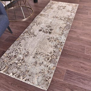 Könnte beinhalten: Ein rechteckiger Teppich mit verblichenem Blumendesign in Grau-, Beige- und Brauntönen. Der Teppich liegt auf einem dunklen Holzboden. Ein blauer Sessel und ein goldener Beistelltisch sind im Hintergrund zu sehen.