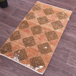 May include: A rectangular area rug featuring a peach-colored background with a repeating pattern of dark brown diamond shapes. Each diamond contains a detailed floral motif in shades of brown and white. The rug has short white fringe on both ends.