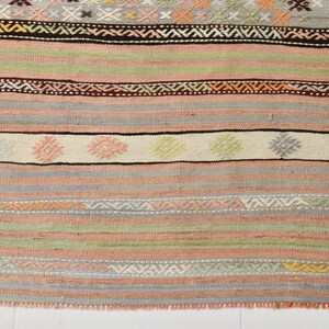 May include: A woven rug with a colorful striped pattern. The rug has a variety of colors, including pink, green, brown, and white. The rug is made of wool and has a fringe on the edges.