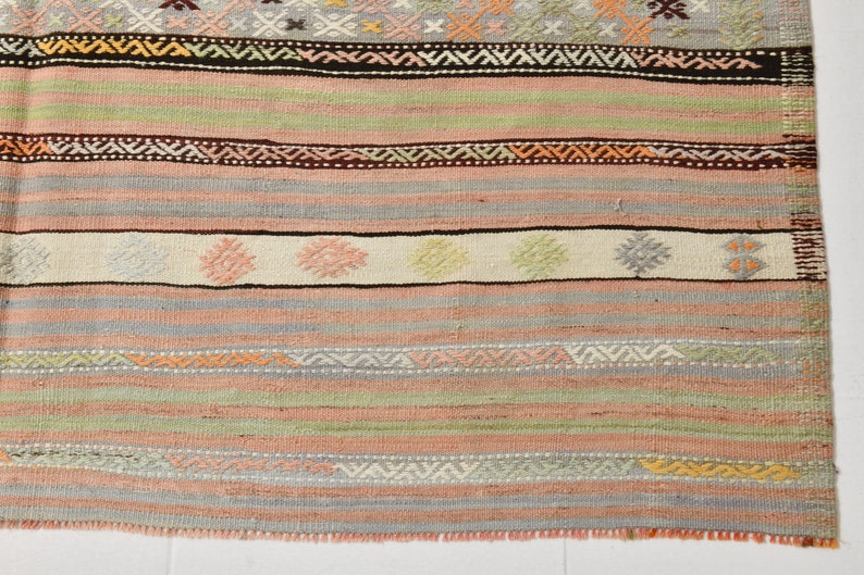 May include: A woven rug with a colorful striped pattern. The rug has a variety of colors, including pink, green, brown, and white. The rug is made of wool and has a fringe on the edges.