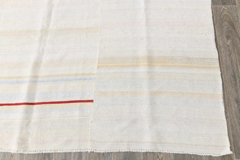 May include: Cream-coloured rug featuring horizontal stripes in beige, light blue, and a bold red accent. The rug has a textured surface and a simple, minimalist design, suitable for various interior styles.