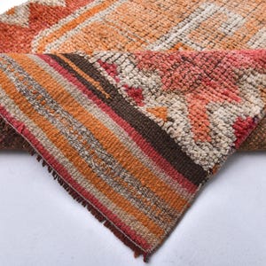 May include: Close-up of a woven rug with a geometric pattern in shades of orange, brown, and pink. The rug has a fringed edge and is folded in half.