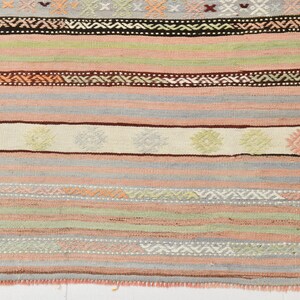 May include: A close-up of a woven rug with a striped pattern in shades of pink, green, brown, and white. The rug has a fringe border.